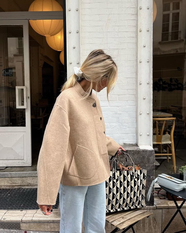 Woman in stylish beige coat with a patterned tote bag stands near a café entrance.