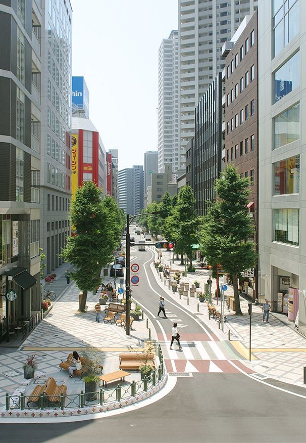 Modern urban street with tall buildings, green trees, and pedestrians walking on a sunny day.