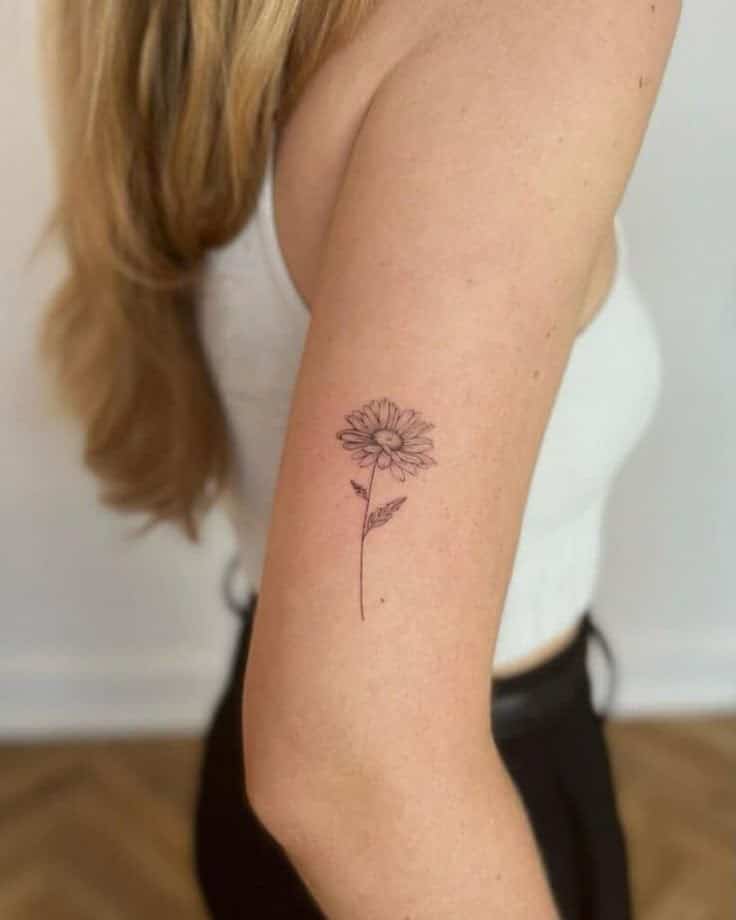 Minimalist daisy tattoo on upper arm, worn by person in white top and black pants. Simple floral body art design.