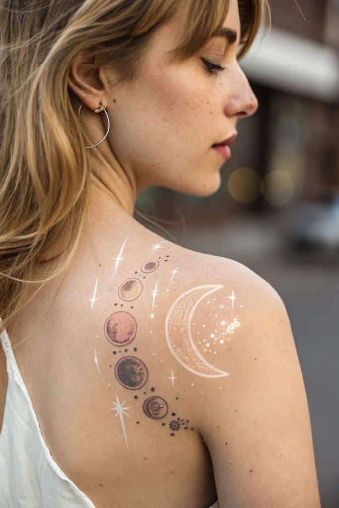 Woman with celestial tattoo on shoulder featuring moon and planets, wearing hoop earrings.