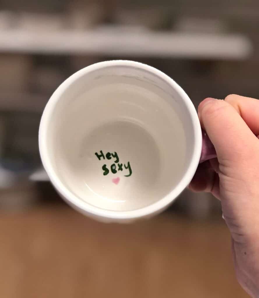 Person holds an empty mug with Hey sexy written inside next to a small pink heart.