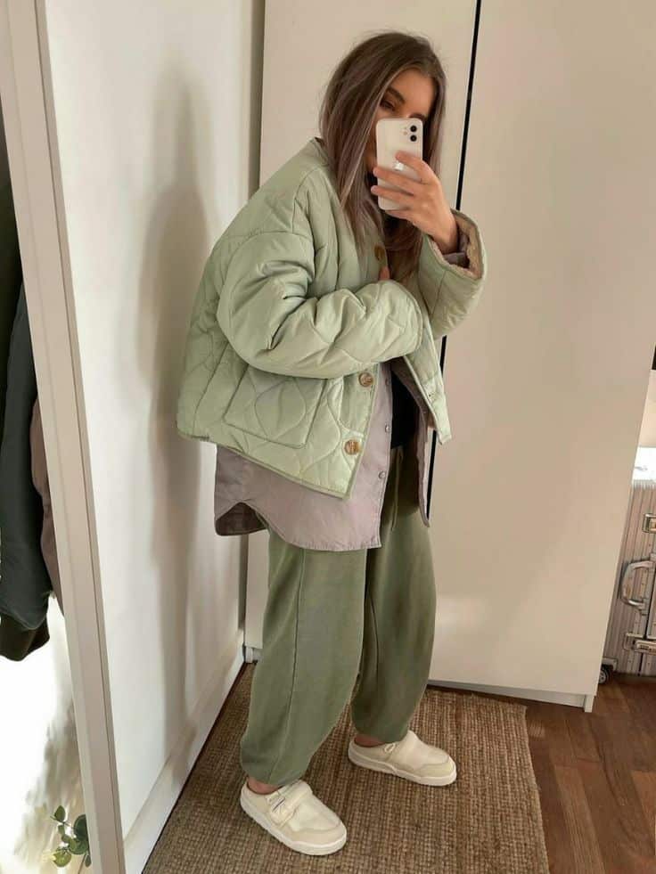 Woman in green quilted jacket and sweatpants takes a mirror selfie in cozy room.