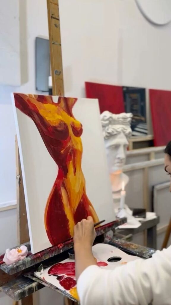 Artist painting vibrant female silhouette on canvas in studio, with palettes and sculptures in the background.