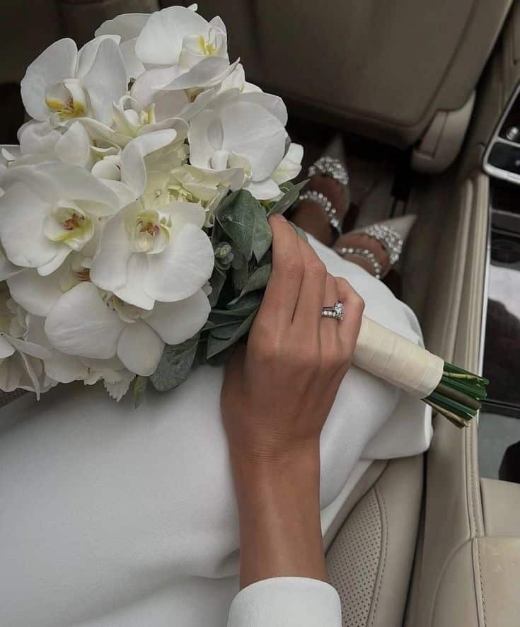 Bridal hand holds white orchid bouquet, displaying rings against a beige car interior. Elegant shoes in the background.