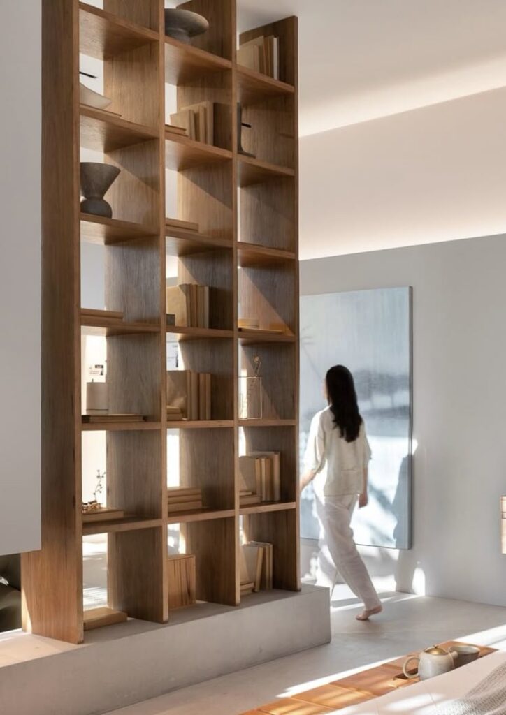 Modern living room with wooden bookshelf and a person standing by a large window, creating a serene atmosphere.