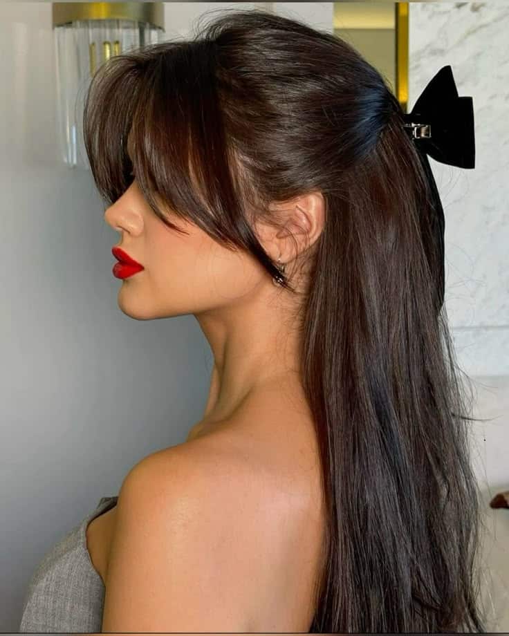 Woman with long, dark hair and red lipstick, side profile. Wearing a black hair bow, against a modern interior background.