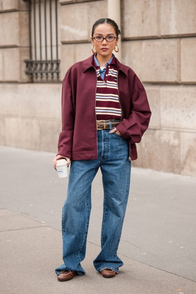 Woman in stylish outfit with maroon jacket, striped shirt, and jeans, holding a coffee cup on city street.