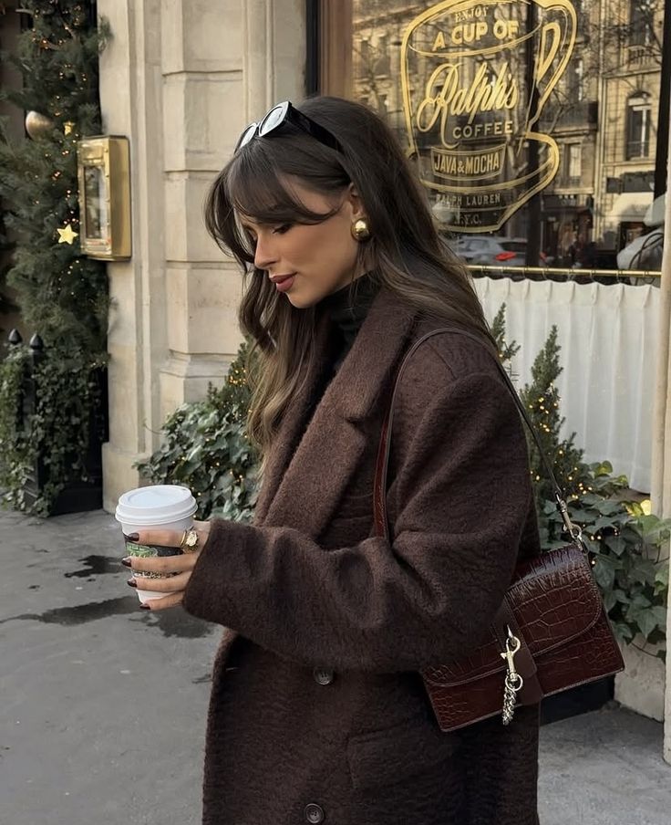 Woman in brown coat holding coffee outside Ralph's Coffee shop, wearing sunglasses and chic handbag.