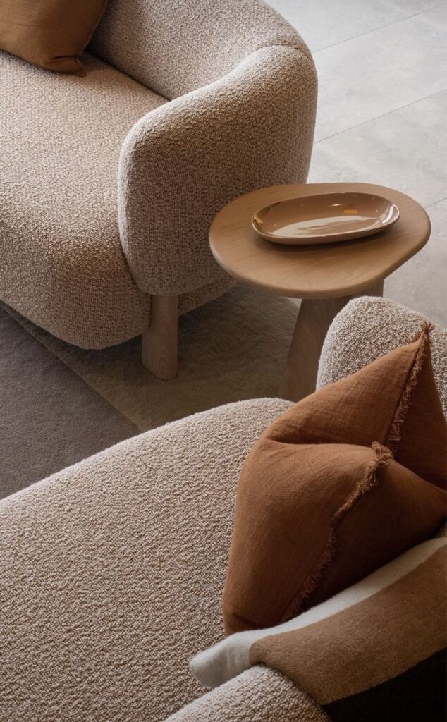 Beige textured sofa with rust pillows, wooden side table, and ceramic dish in a modern living room setting.