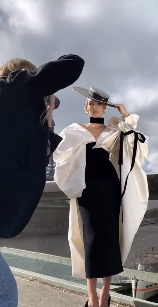 Fashion photoshoot: model in elegant dress and hat, photographer in action against cloudy sky backdrop.