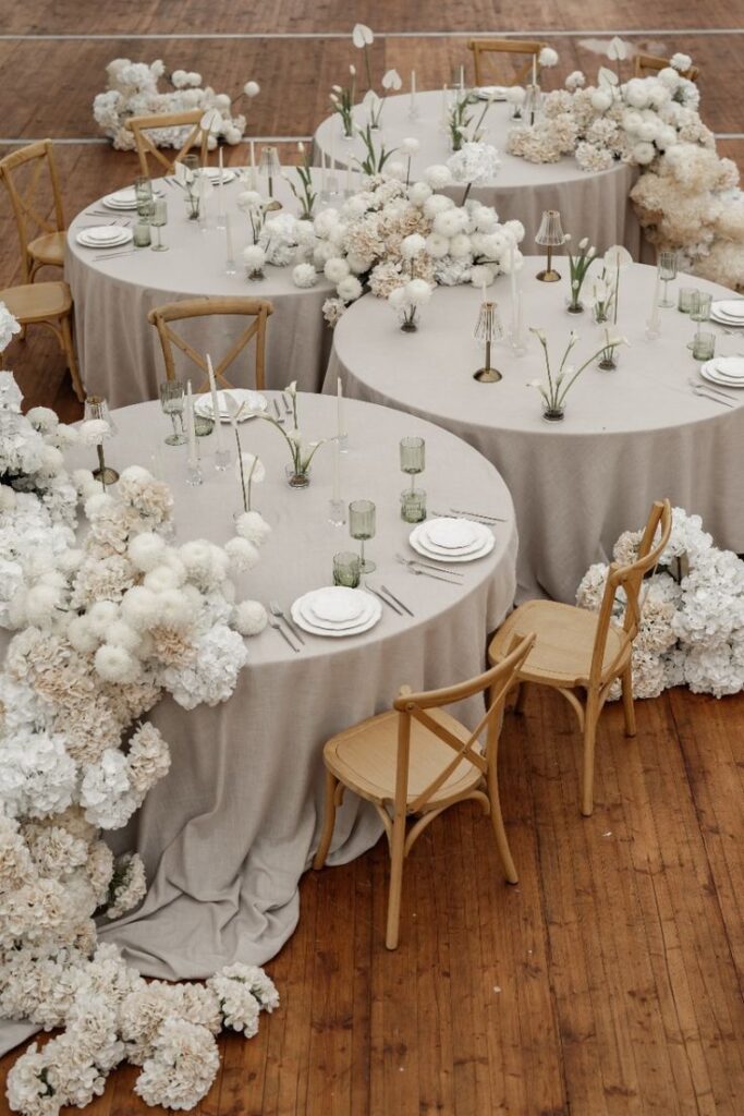 Elegant wedding tablescape with white floral arrangements, wooden chairs, and beige linens on a wooden floor.