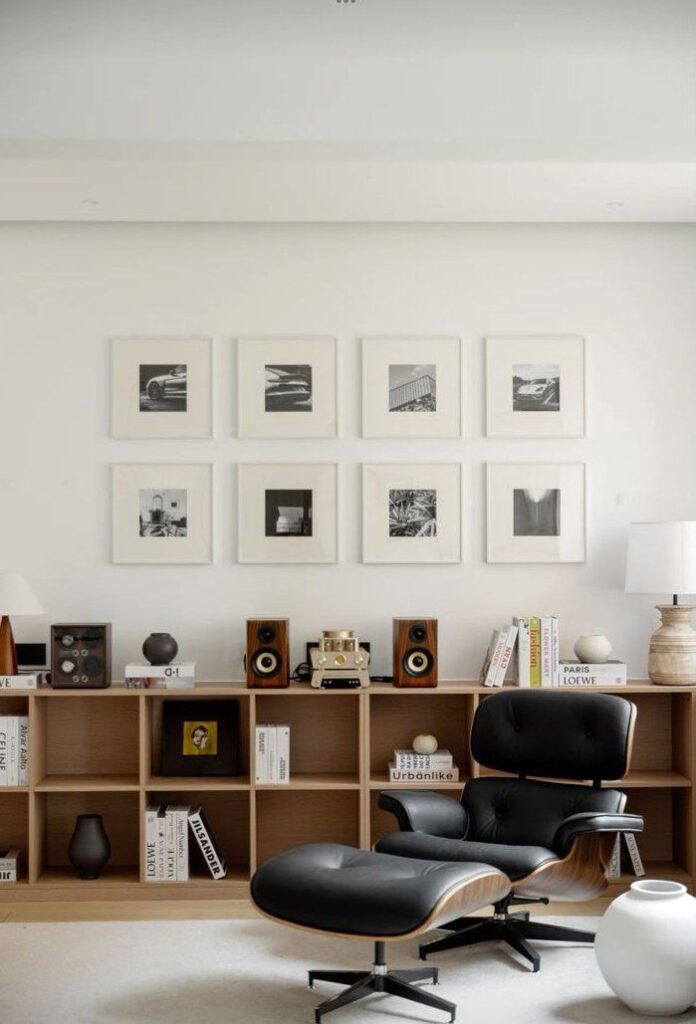 Modern living room with black lounge chair, bookshelf, art display, and speakers. Minimalist decor and neutral tones.