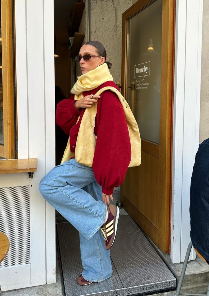 Fashionable woman in red jacket and jeans posing at cafe door, wearing sunglasses and stylish scarf.