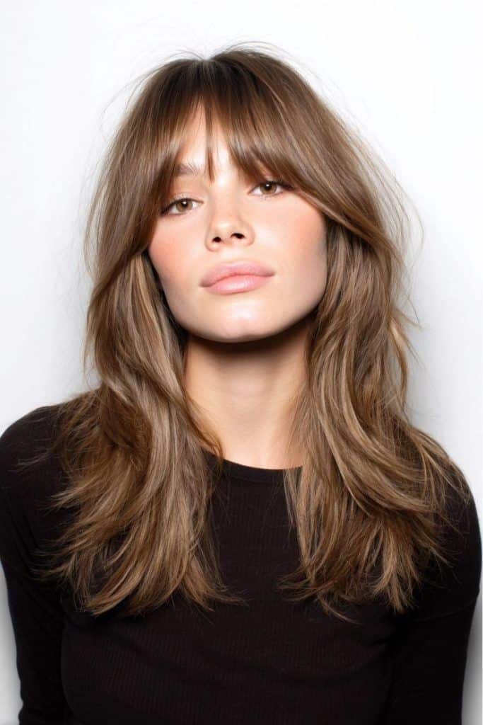Woman with layered brown hair and bangs, wearing a black shirt, against a white background.
