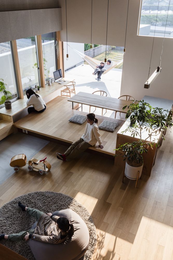 Modern open-concept living room with people relaxing, hammock visible outside, wooden floors, and natural light.