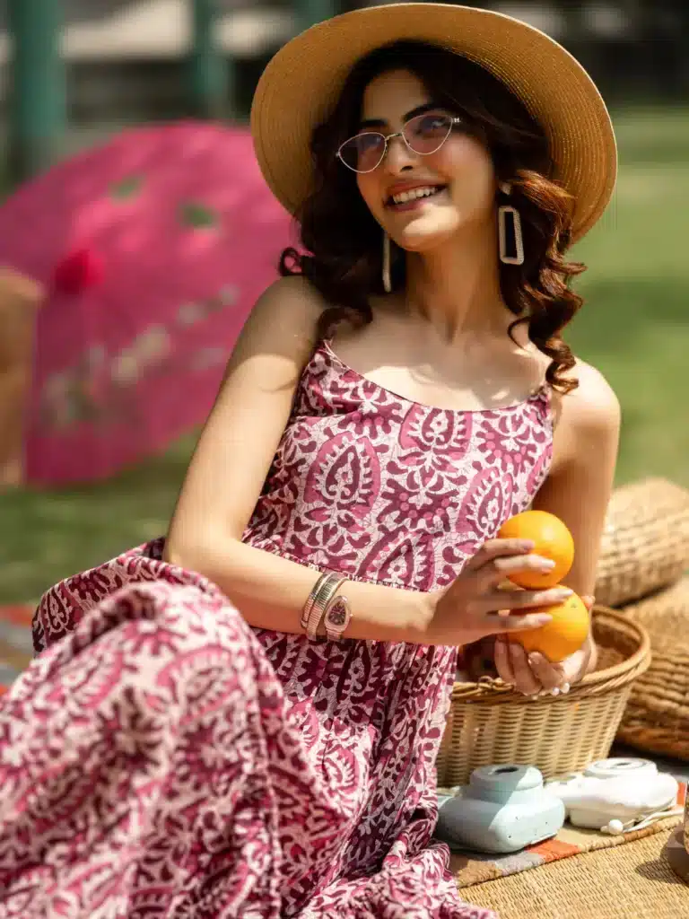 Woman in patterned dress and hat holds oranges at a picnic, surrounded by baskets and a pink parasol.