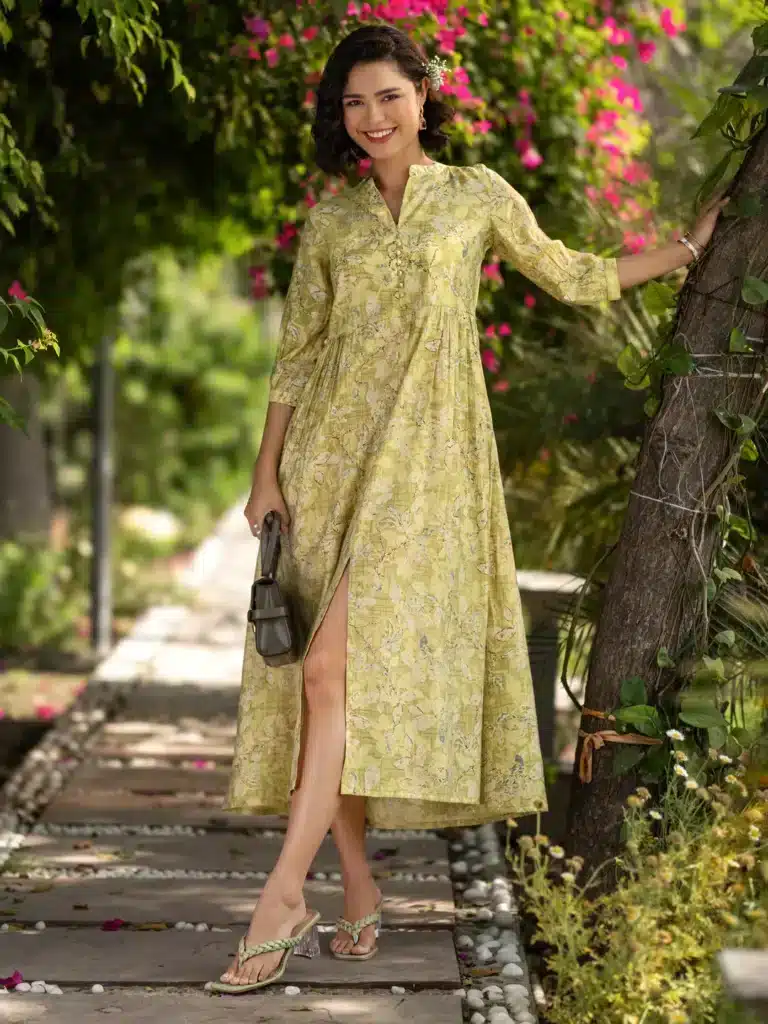 Woman in a floral yellow dress smiling, standing outdoors near a tree with colorful flowers.