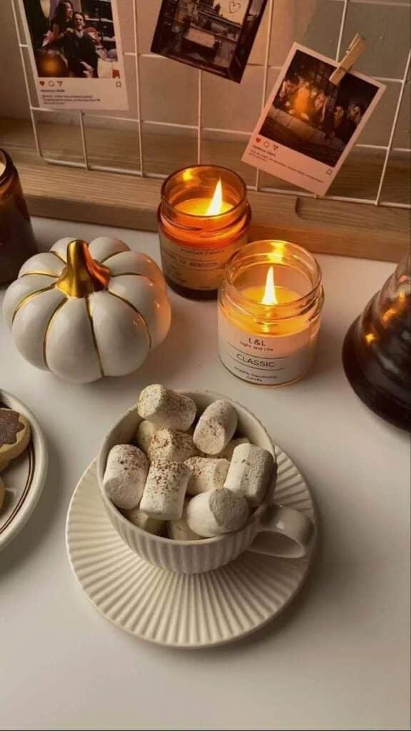 Cozy autumn vibes with hot cocoa, marshmallows, lit candles, and a decorative pumpkin on a table.
