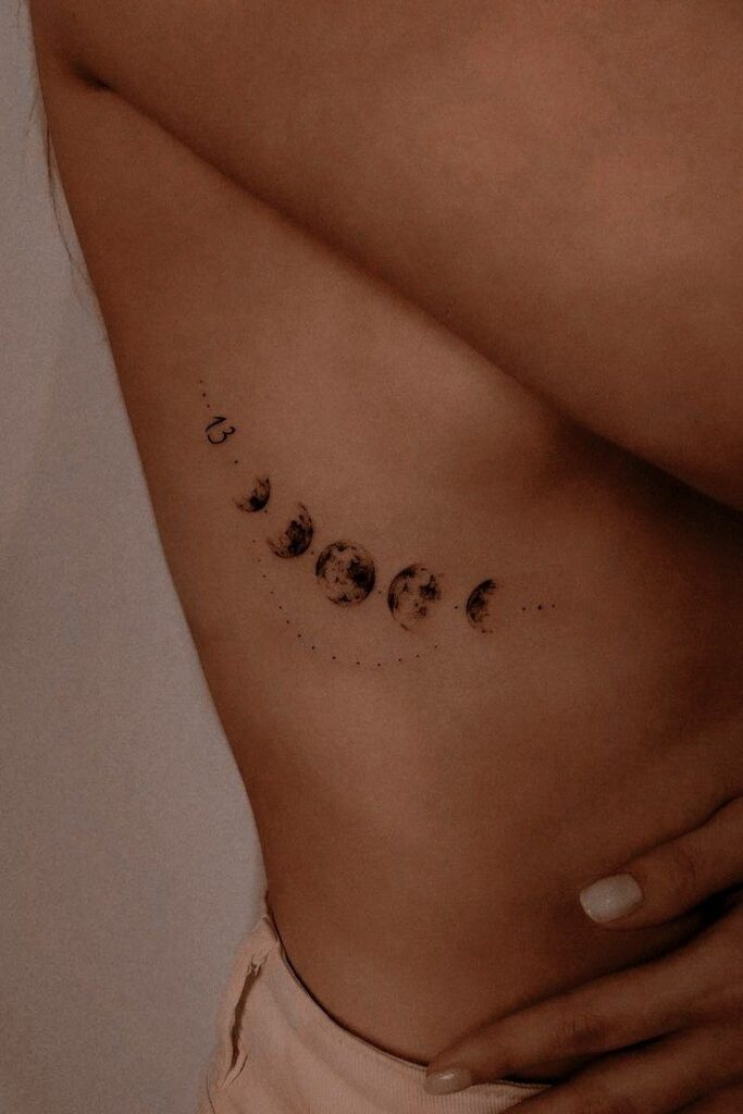 Moon phases tattoo on ribcage, delicate design featuring lunar cycle, personal body art against tan skin.