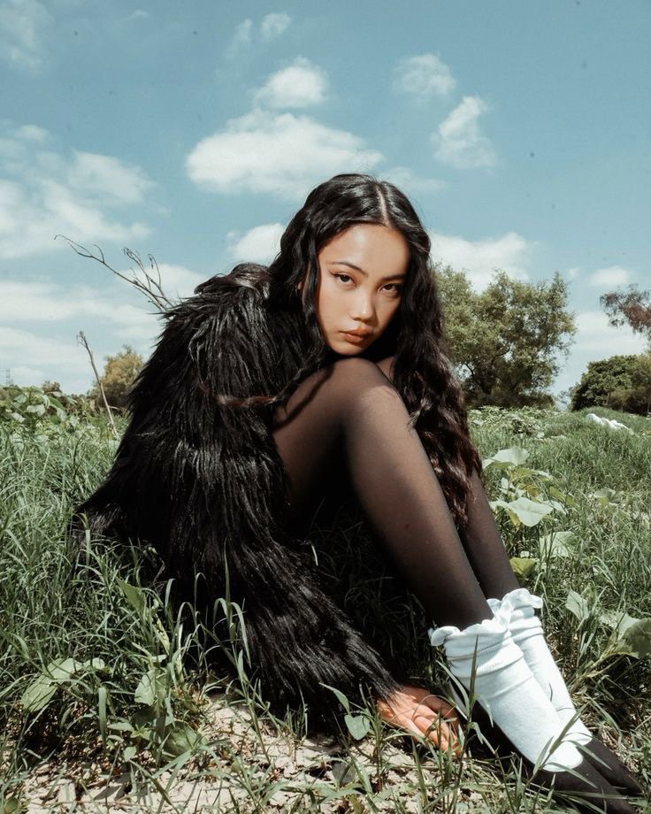 Young woman in black fur coat sitting in a grassy field under a blue sky.
