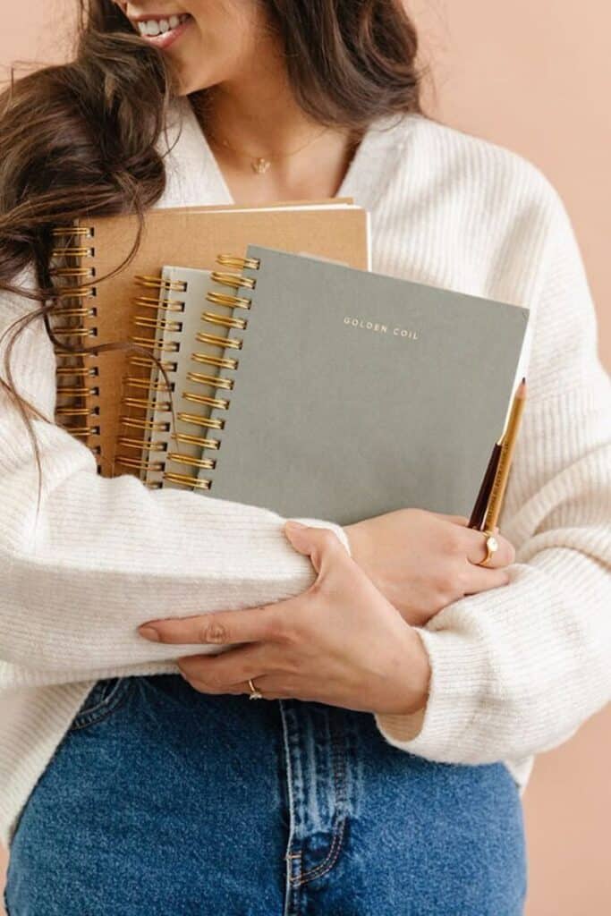 Woman holding spiral notebooks and a pen, wearing a cozy sweater and jeans, with a neutral background.