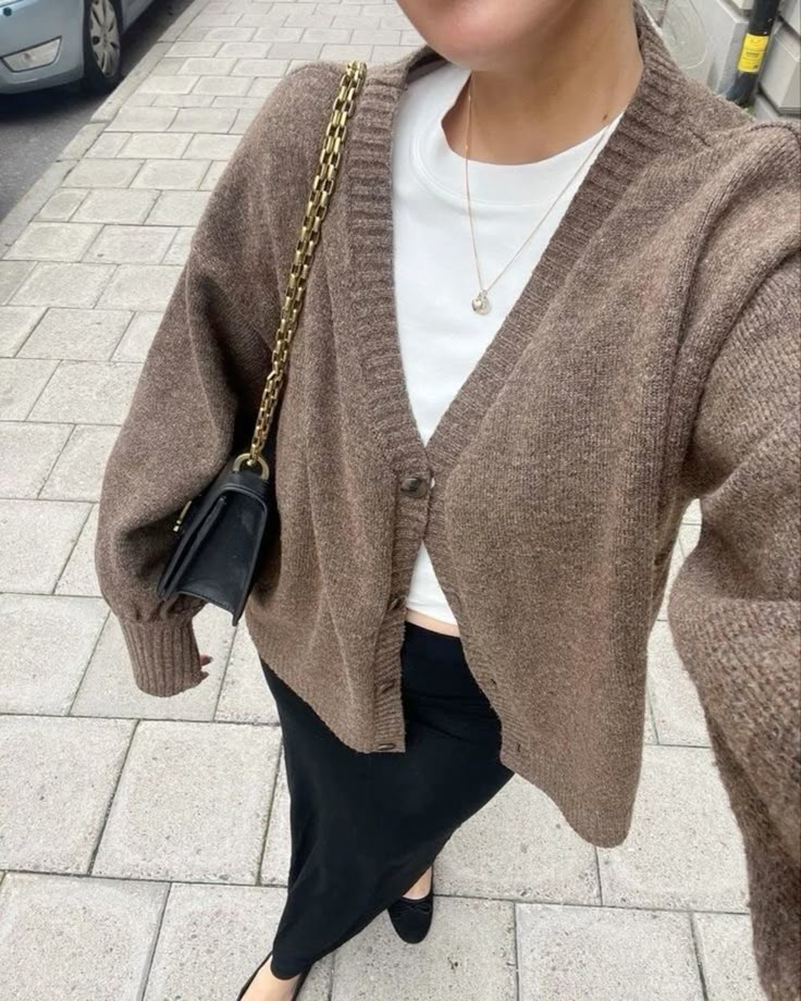 Woman in brown cardigan, white shirt, and black skirt walking with a handbag on a city sidewalk. Fashionable street style.