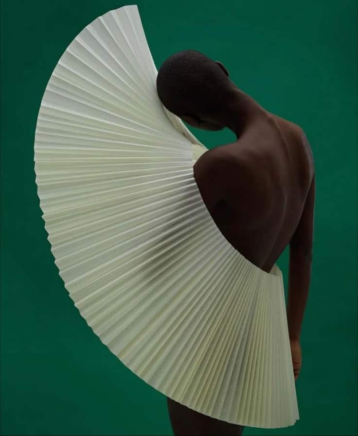 Person posing with an artistic, pleated structure against a green background, showcasing fashion and creativity.
