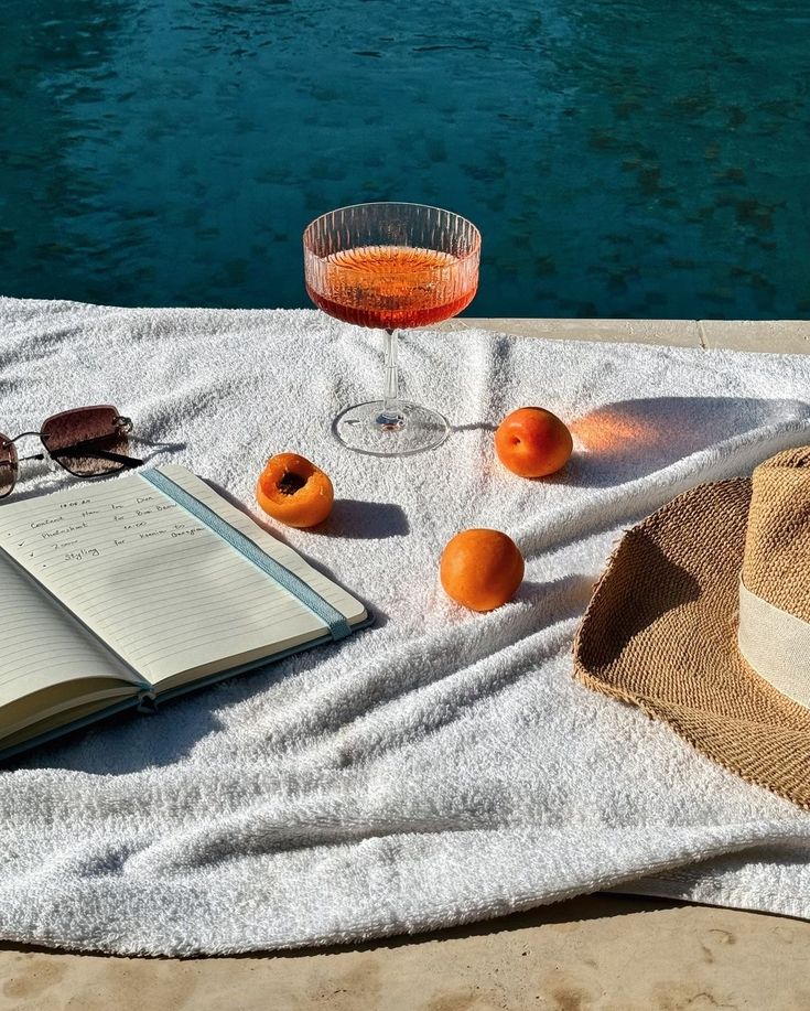 Summer poolside scene with a drink, apricots, a notebook, sunglasses, and a hat on a towel.