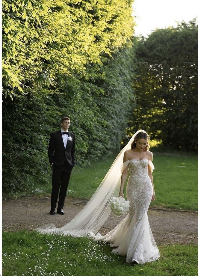 Bride in elegant lace gown with train and bouquet; groom in tuxedo in lush garden setting.