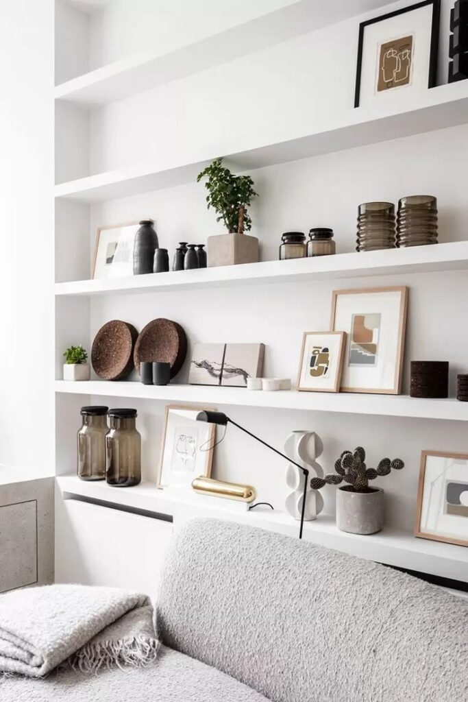 Modern living room with white shelves, decorative vases, art frames, and a cozy gray couch with blankets.
