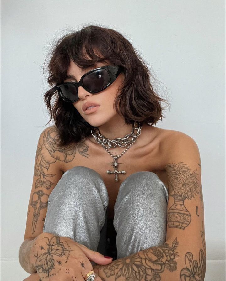 Tattooed woman in sunglasses and silver pants, wearing a chain necklace, poses against a plain background.