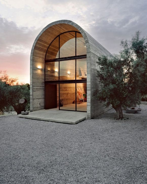 Modern arched concrete house with large glass facade surrounded by trees at sunset.