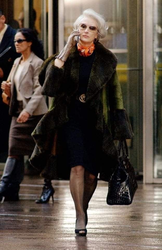 Stylish woman in fur coat and sunglasses walking while talking on phone, holding a black bag. Elegant urban fashion.