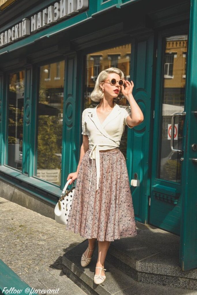 Vintage fashion woman in skirt and sunglasses stands outside a teal store, holding a white purse.