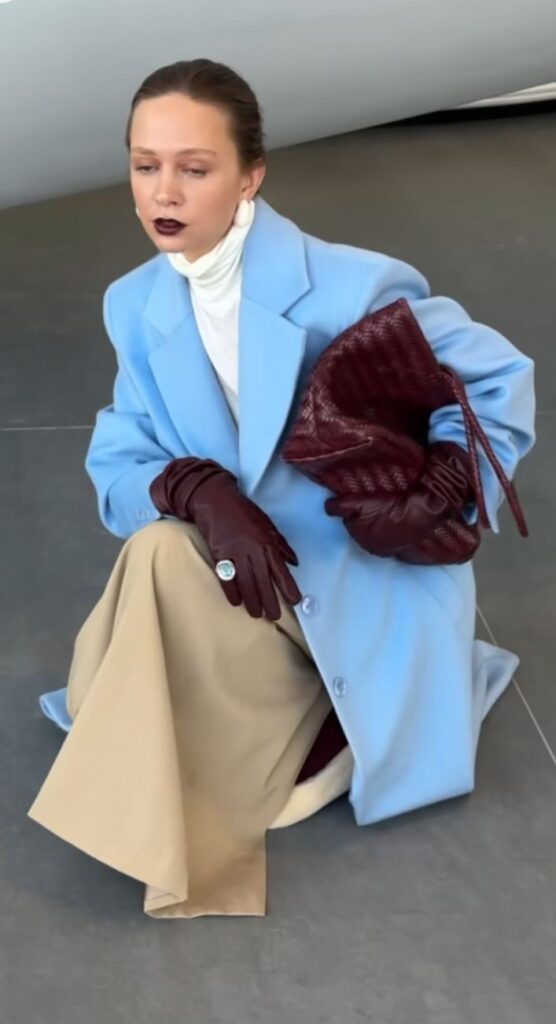 Fashionable woman in a light blue coat, burgundy gloves, and beige skirt holding a textured bag.