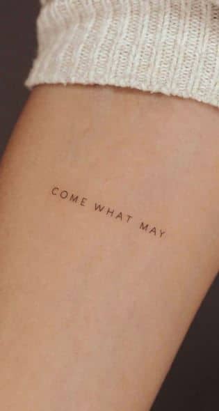 Minimalist arm tattoo reading Come What May in simple font, on skin with a light knit sleeve partially visible.