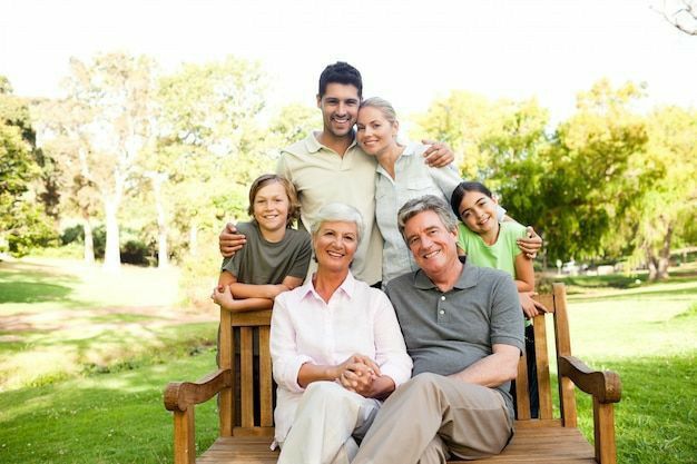 Happy multigenerational family smiling outdoors, sitting and standing on a park bench with a lush green background.
