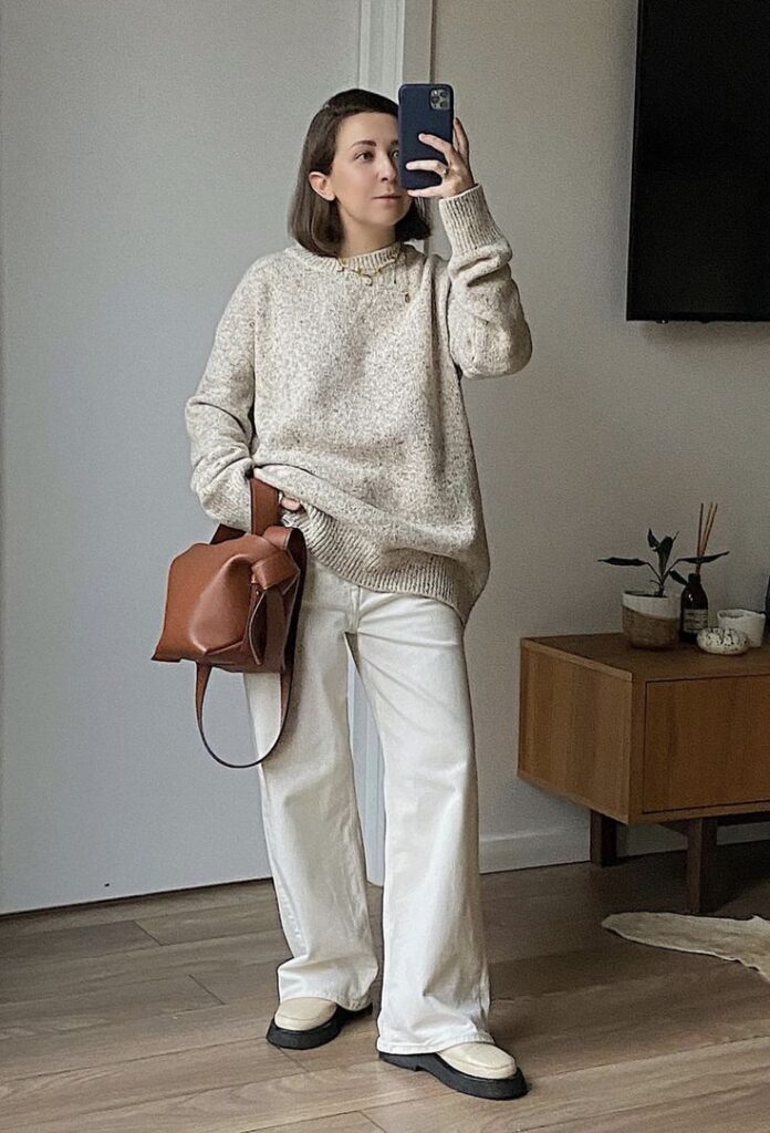 Woman in cozy sweater and trousers takes mirror selfie, holding brown bag, showcasing casual chic style at home.