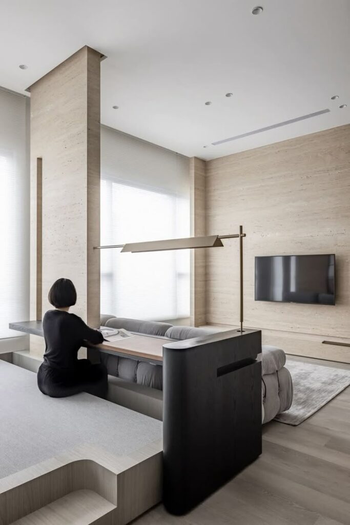 Modern minimalist interior with woman sitting at built-in table, neutral tones, and wall-mounted TV.
