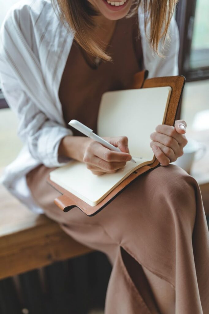 Person writing in a journal with a pen, seated beside a window.