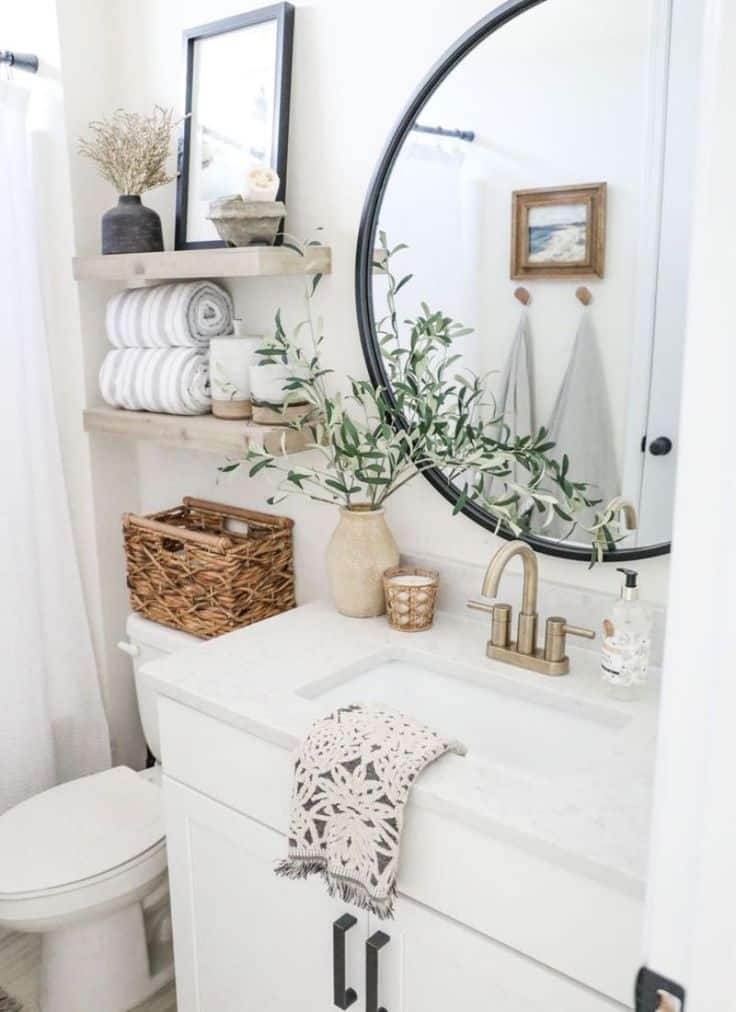 Chic bathroom with round mirror, white vanity, brass faucet, woven basket, and decorative shelves with greenery accents.