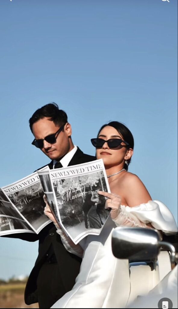 Newlywed couple in sunglasses reads Newlywed Time newspaper while sitting on a scooter under a clear blue sky.