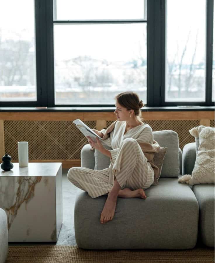 Woman in cozy loungewear reading on a gray couch by a large window, with a marble table and a cup beside her.