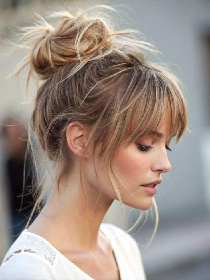 Woman with blonde messy bun hairstyle and bangs wearing a white top, side profile view.