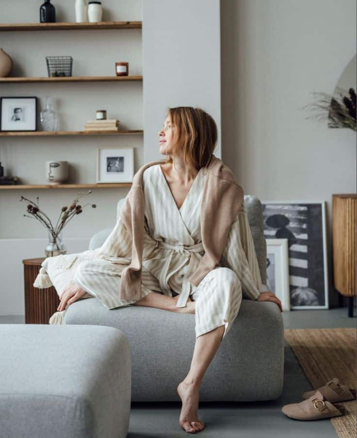 Woman in a cozy robe sits on a sofa in a stylish living room with shelves and decor elements.