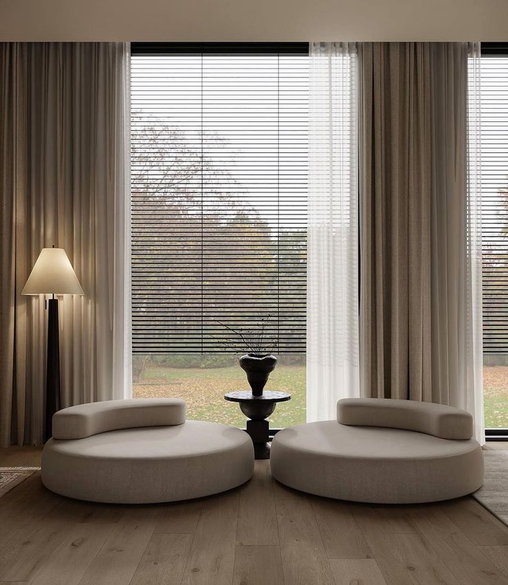Modern living room with round sofas, large window, floor lamp, and neutral curtains. Stylish and minimalist interior design.