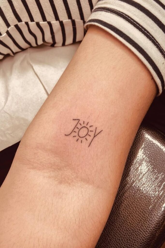 Minimalist 'Joy' tattoo on wrist with sun illustration, symbolizing happiness and positivity close-up.
