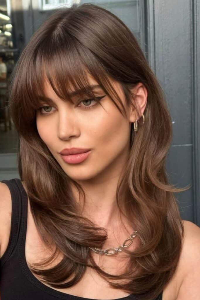 Woman with layered brunette hair and bangs, wearing a black top and chain necklace.