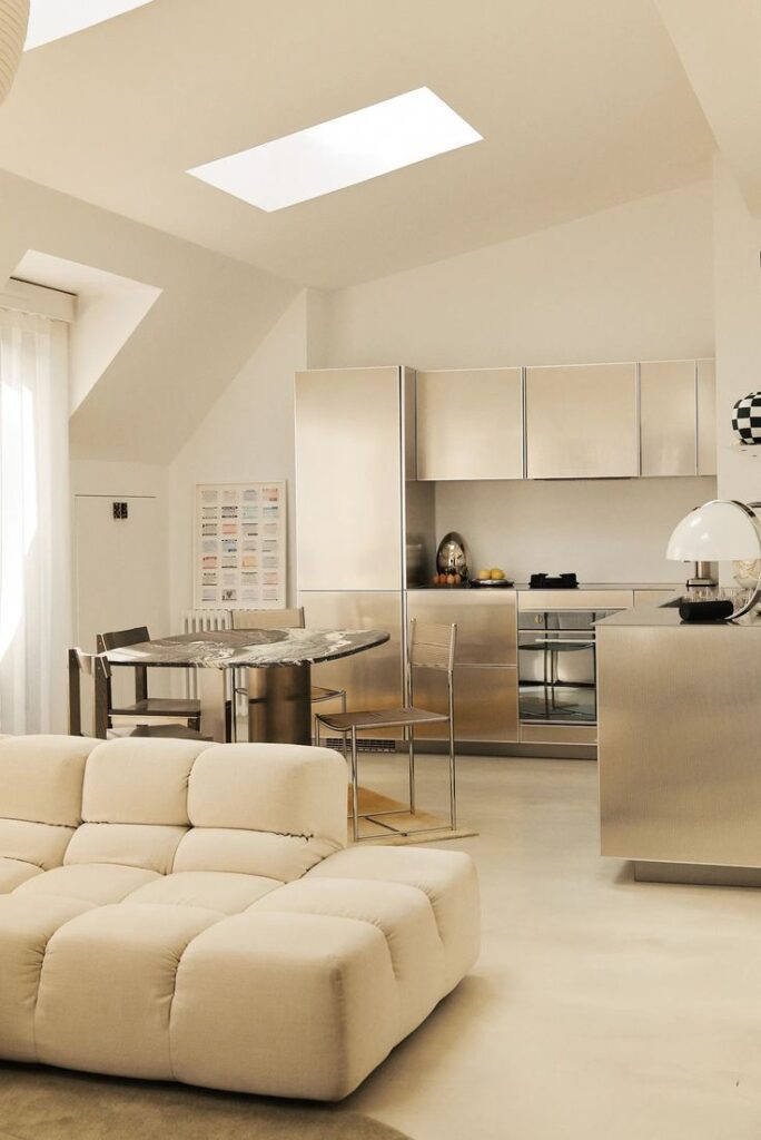 Modern kitchen with stainless steel cabinets, marble table, and beige sofa in bright loft apartment.