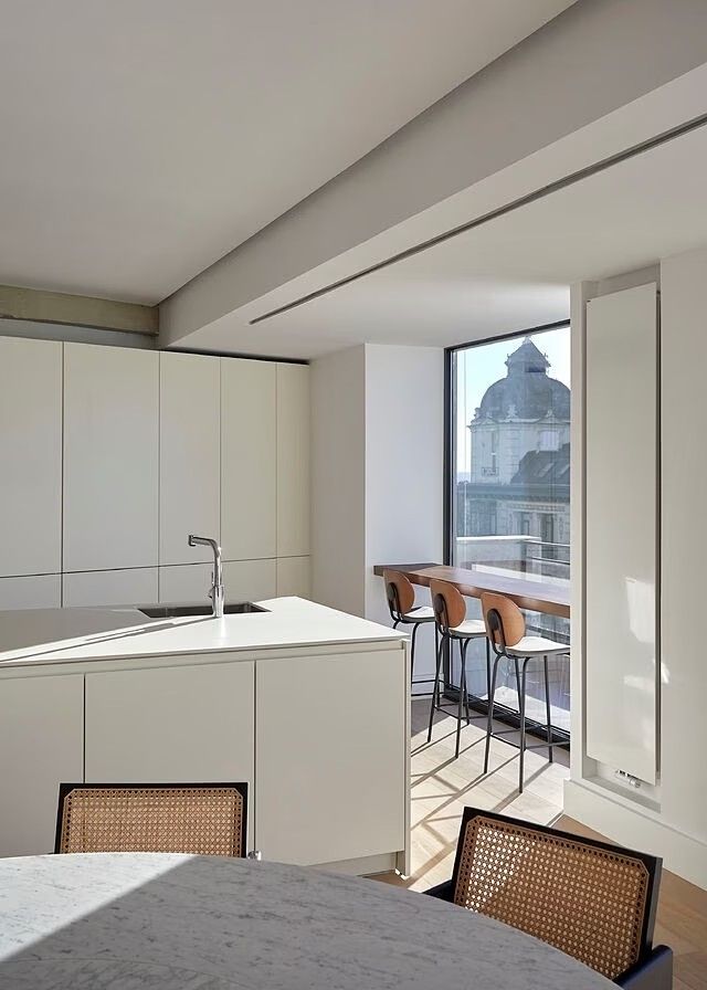 Modern kitchen with white cabinets, island sink, round table, and city view through a large window. Sleek and minimal design.